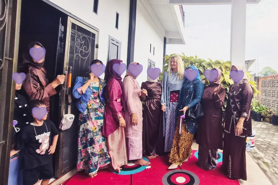 Sepuluh perempuan dan anak memakai pakaian adat berbaris di beranda bersama Tine. Semua wajah kecuali wajahnya ditutup hati ungu.