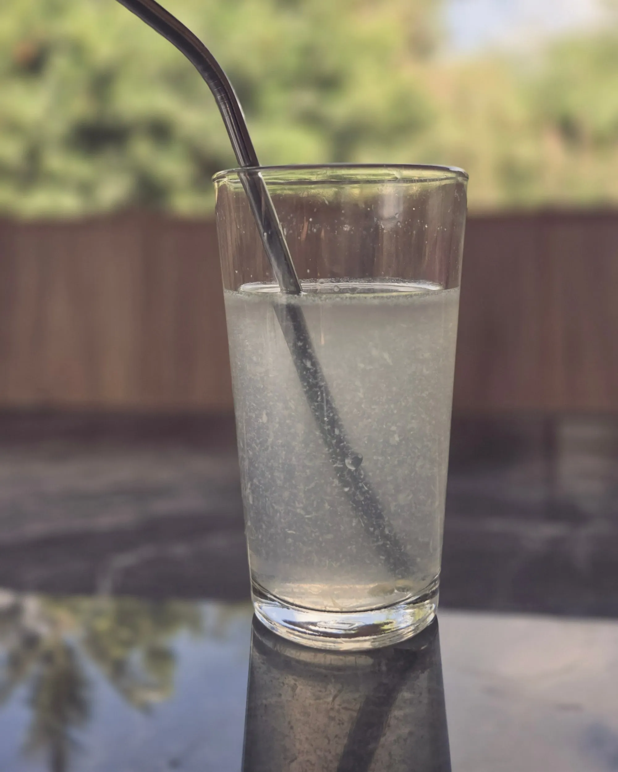 Minuman yang sudah jadi di gelas tinggi dengan sedotan logam, terpantul di meja gelap, dengan pepohonan hijau di belakang.