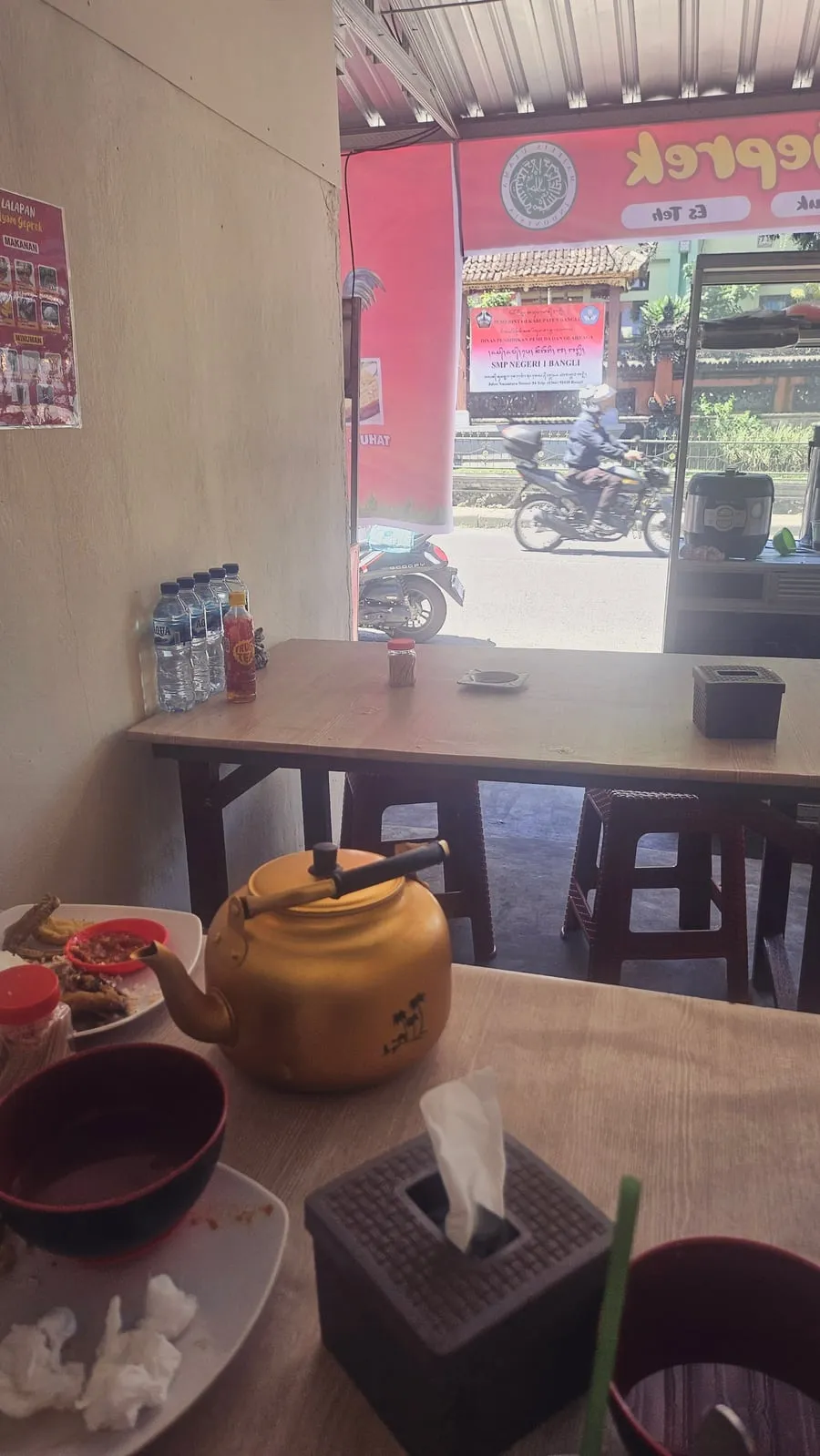 Teko keramik di atas meja kayu di kafe terbuka, dengan jalan di luar.