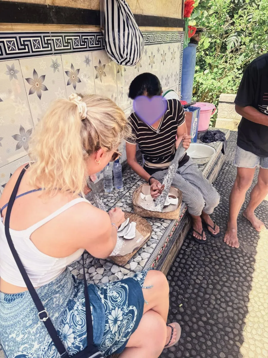 Seorang ibu memarut kelapa dengan pisau di atas keranjang, sementara Tine duduk di sampingnya dan membantu. Wajah ibu itu ditutup hati ungu.
