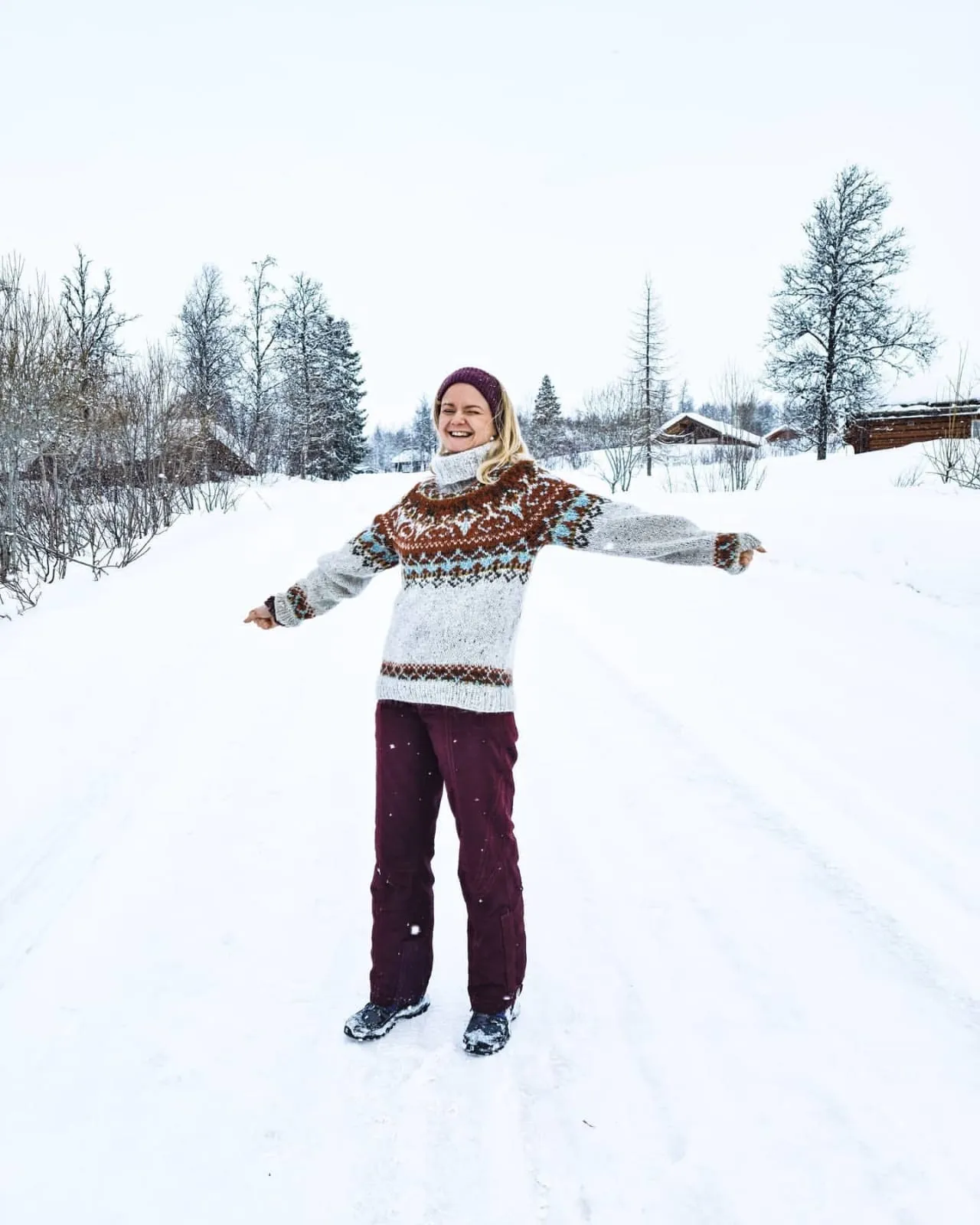 Tine berdiri di tengah jalan bersalju di Norwegia dengan tangan terentang, memakai sweter wol bermotif.