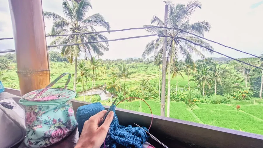 Dua tangan merajut sesuatu berwarna biru di jarum bundar, dengan pohon kelapa dan sawah hijau di balik pagar.