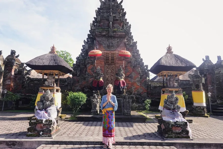 Tine memakai sarung di depan gerbang pura dengan payung merah dan patung batu.